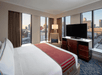 Hotel room with city view, featuring a king-sized bed, a television on a wooden stand, a cushioned chair with a lamp, and large windows with curtains.