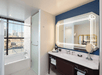 Modern bathroom with a white bathtub, frosted glass shower door, large illuminated mirror above a double sink vanity, and a view of city buildings through a window.