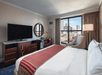 Hotel room with a large bed, red throw, and a circular mirror above a desk. A TV is on a stand opposite the bed. A window with city views is beside an armchair and lamp.