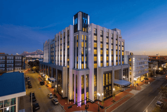 A modern, multi-story hotel building at dusk with illuminated signs and vertical lighting accents, situated on a city street corner.