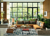 Modern living room with large windows, an L-shaped orange sofa, indoor plants, patterned rug, and mid-century style furniture. Natural light fills the space.