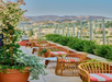 Outdoor rooftop seating area with wicker chairs and small tables, potted plants, glass railing, and a view of the Hollywood Hills and Hollywood sign in the distance.