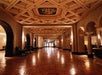 A grand hallway with a patterned ceiling, arched doorways, and polished tile flooring, featuring seating areas along the sides.
