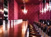 A long bar with empty glasses and chairs lined up on one side. The room features red patterned wallpaper and chandeliers.
