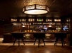 Dimly lit bar with three stools, a wooden counter, and shelves stocked with bottles. A bartender prepares a drink in front of a mirror.