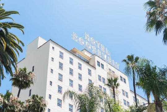 Exterior view of Hotel Roosevelt, a white multi-story building with a neon sign on top, surrounded by palm trees under a clear blue sky.