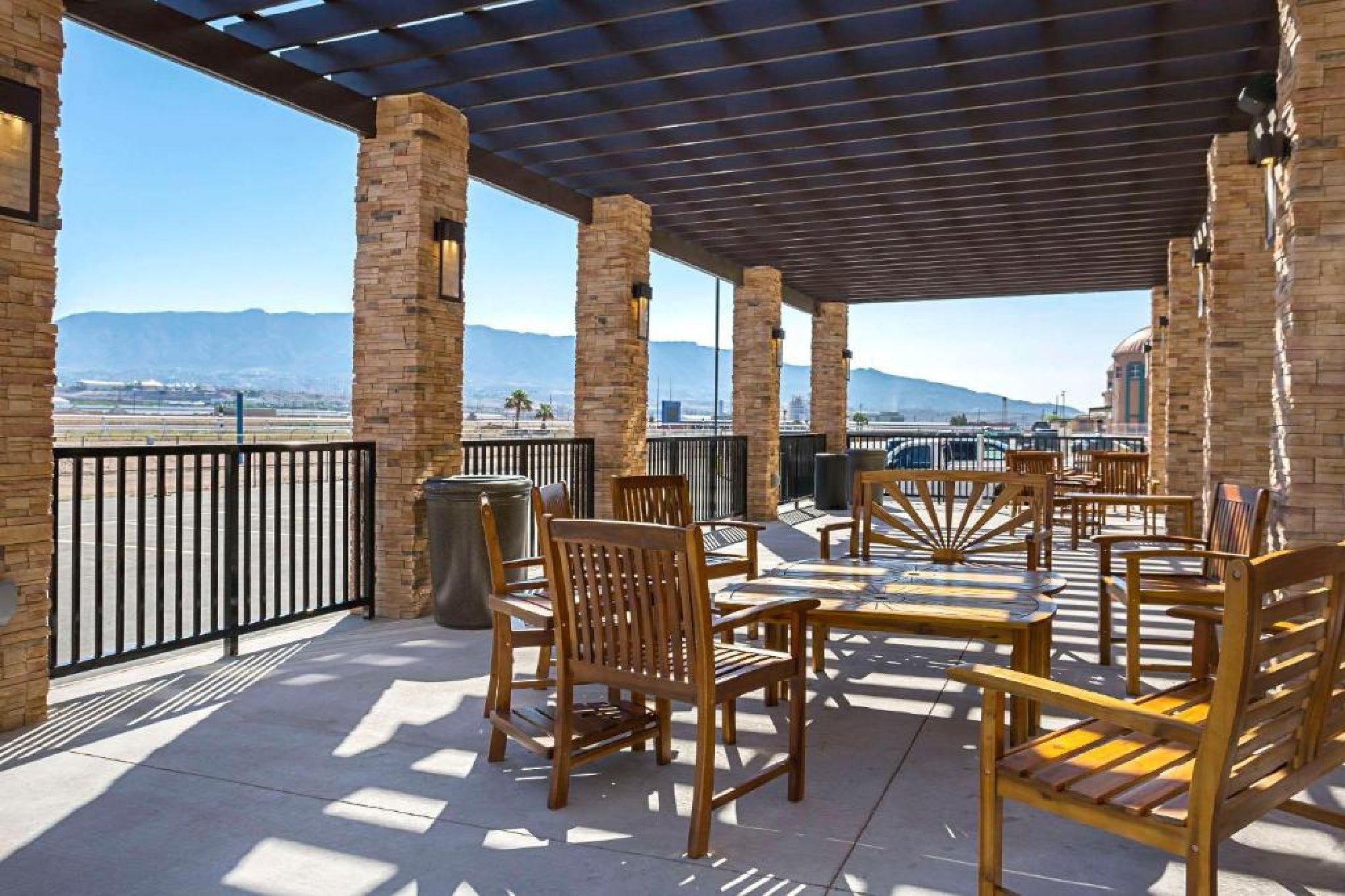 Outdoor patio with wooden tables and chairs under a pergola, stone columns, and a view of mountains and a fence in the background.