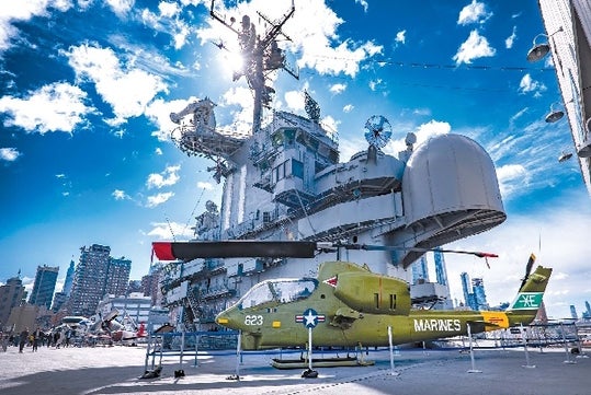 A olive green military plane on the flight deck with other military equipment behind it on a bright sunny day in New York City.