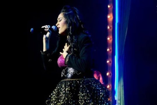 A woman sings on stage, holding a microphone with one hand and placing the other on her chest. She wears a sparkly dress and dark jacket. Bright lights illuminate the background.