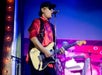 Musician in a red shirt and sunglasses plays electric guitar and sings into a microphone on a colorful stage.