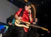 Person wearing a red outfit and sunglasses plays an electric guitar on stage with other performers.