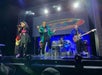 Band performing on stage, with a singer, guitarist, and drummer. Bright lights illuminate the scene, and a large screen displays colorful graphics in the background.