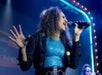 A woman sings passionately into a microphone on stage, wearing a sparkly blue top and a dark jacket.