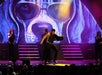 A performer in a gold jacket dances on stage with backup singers, in front of a large screen displaying an image of a dog wearing sunglasses.