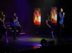 Four performers in blue outfits sing on a dimly lit stage with palm tree sunset backgrounds projected behind them.