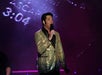 A man in a gold sequin jacket sings into a microphone on stage, with a digital clock display reading "3:04" in the background.