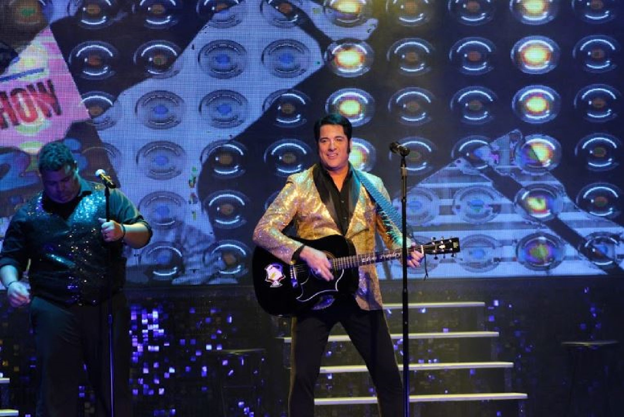 A performer in a gold jacket plays a black acoustic guitar and sings on stage with another musician and a backdrop of illuminated circular patterns.
