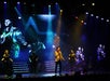 A performer in a gold jacket sings on stage with backup singers and a musician, while black-and-white images of Elvis Presley display on a large screen behind them.