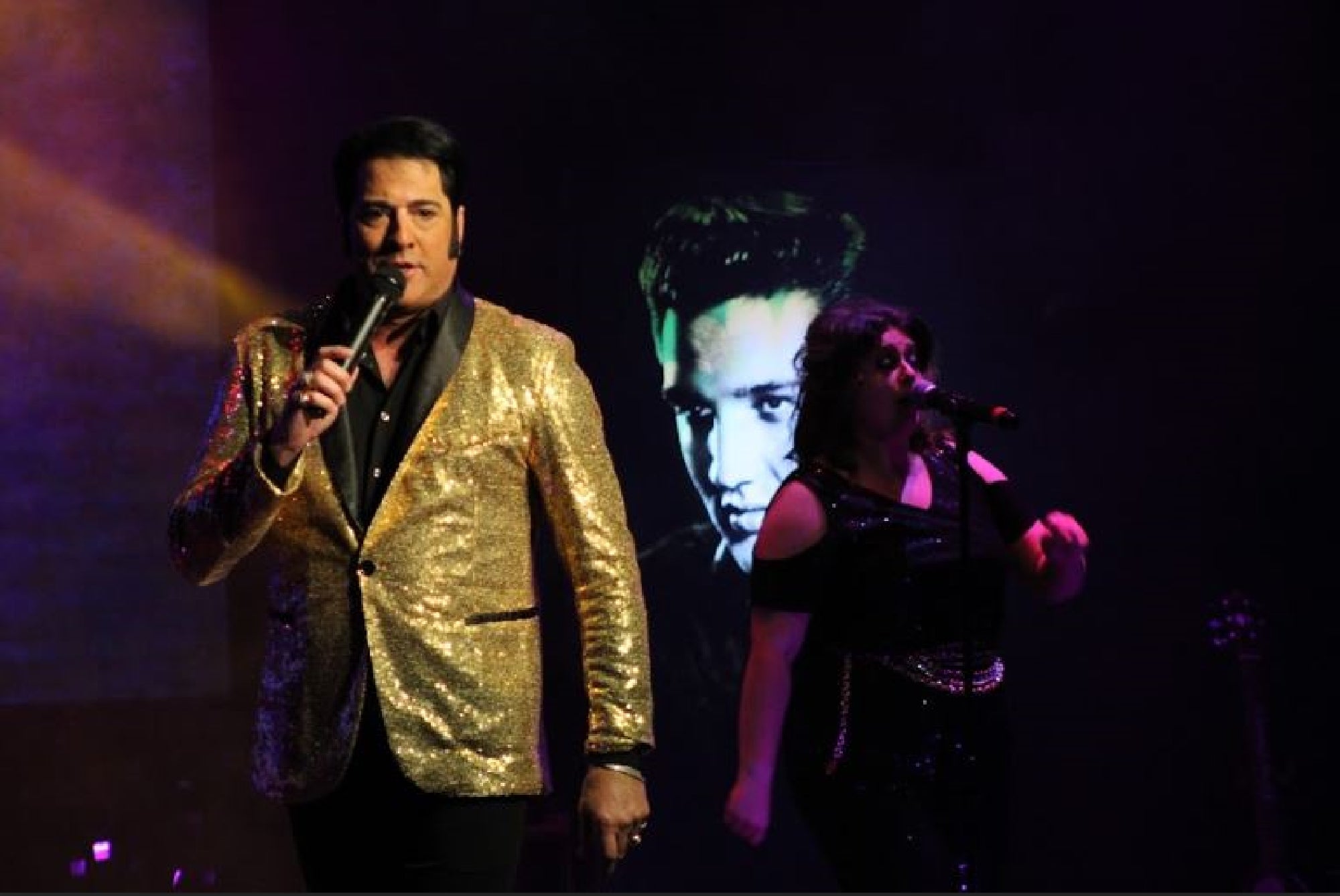 A male singer in a gold jacket performs on stage with a female singer, while a black-and-white image of another man is displayed in the background.