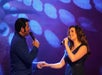 A man and a woman are holding hands and singing into microphones on stage with a purple and blue lit background.