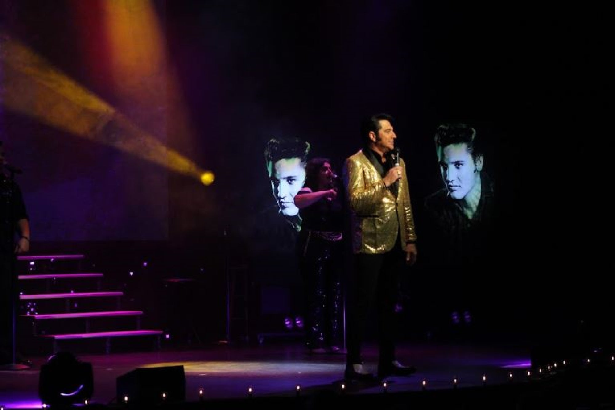 A man in a gold jacket sings on stage with projected images of Elvis Presley behind him and colorful stage lighting.