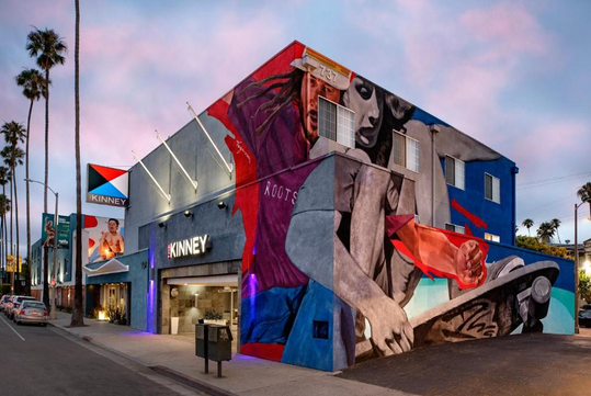 Colorful mural on a modern two-story building, featuring abstract figures and a skateboard. Palm trees line the street, and a sign reads "Kinney" near the entrance.