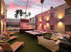 Outdoor lounge area with string lights, ping pong table, fire pit, and seating. Palm trees and building exterior with illuminated signs in the background. Twilight setting.