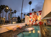 An outdoor pool area at dusk, with lounge chairs and palm trees. A large mural of a man in swim trunks is on the building wall. City buildings and billboards are visible in the background.