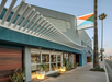Modern building exterior with large windows, overhang, and "Kinney" sign. Sidewalk with potted plants and a tall palm tree in the background. Sky is clear.