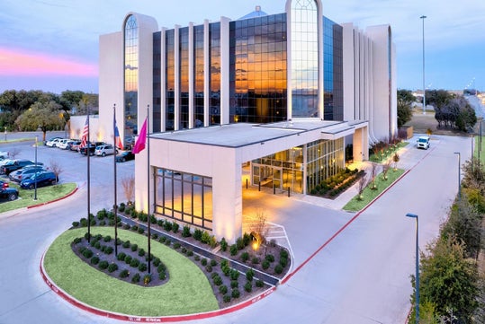 Modern office building with glass windows reflecting sunset, multiple floors, landscaped entrance, and parking lot with cars and flagpoles in front.