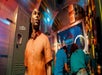 A person in an orange shirt stands in a colorful room with a "DESCEND" sign. Two people in blue attire work at a station in the background.