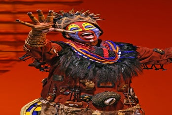 Performer in colorful face paint and elaborate costume sings on stage against an orange background, wearing beads, feathers, and tribal-inspired accessories.