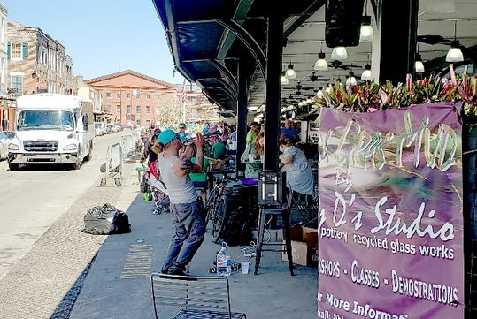 A man takes a photo at an outdoor market with various vendors, a banner promoting a studio, and a truck parked on the street.