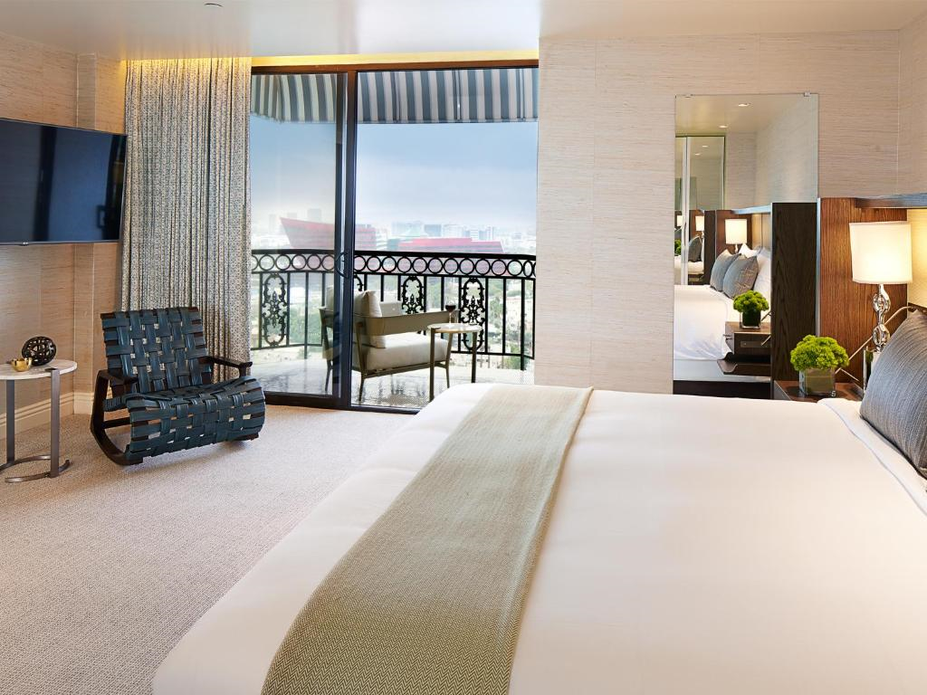 Elegant hotel room with a large bed, armchair, and floor-to-ceiling window leading to a balcony with city views.