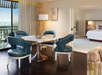 A modern hotel suite with a dining table and four chairs, a bed in an adjoining room, and a balcony with city views.