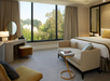 Hotel room with a large window, beige sofa, bed with gold accents, desk with a mirror, and a small round table with a book. Trees are visible outside.