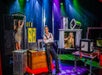 Magician on stage with assistants in sparkly dresses, surrounded by magic props. Colorful lights illuminate the scene.