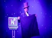 Magician in a top hat performs on stage with a suitcase and a large cloth, set against a backdrop of colored lights.