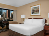 Hotel room with a large bed, bedside tables, lamps, a desk and chair by the window, city skyline visible outside, and a framed painting above the bed.