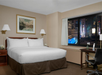 Hotel room with a large bed, two bedside tables, a desk with a chair, and a window showing a cityscape with illuminated buildings at night.