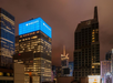Nighttime cityscape featuring illuminated office buildings, including the Barclays building with a bright blue sign, under a cloudy sky.