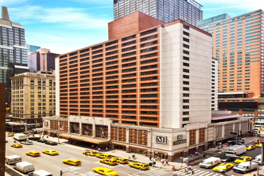 A tall hotel building stands on a busy Manhattan street corner, surrounded by other high-rise buildings and numerous yellow taxis.