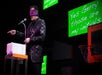 A blindfolded man stands at a microphone and gestures, with bright green screens behind him displaying the text, "Yes Gerry those are my initials.