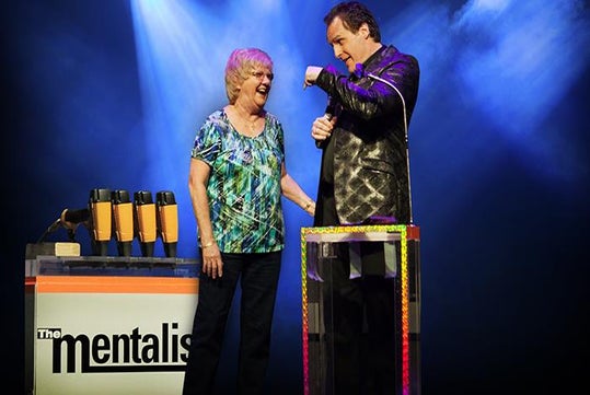 A man in a black suit performing a trick with an older woman in a green shirt next to him on stage at The Mentalist in Las Vegas, NV