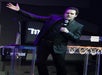 A man wearing a black suit speaking into a microphone with his other arm out on stage at The Mentalist in Las Vegas, NV.