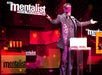 A man in a black suit wearing a red blind fold standing on stage behind a podium with his arms out at The Mentalist in Las Vegas.