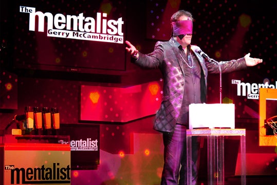 A man in a suit and blindfold stands on stage with arms outstretched, performing as "The Mentalist" with the name Gerry McCambridge displayed in the background.