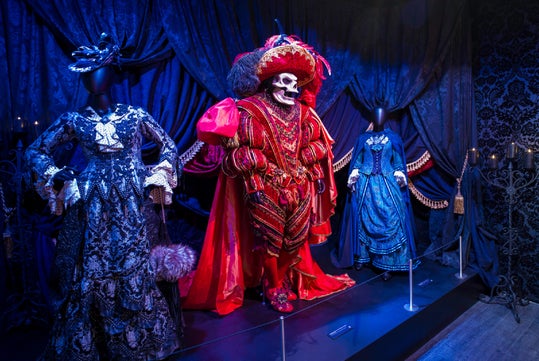Three ornate costumes are displayed on mannequins in a dark, dramatic setting; the central figure wears a red skull mask and elaborate red attire, flanked by two blue and black gowns.