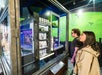 Two people observe a detailed miniature diorama displayed in a glass case at an exhibition, with a green backdrop showing flying dragons.