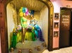 Two mannequins wear colorful feathered showgirl costumes in a decorative archway next to a black door labeled "Stage Door Enquiries." Framed photos hang on the pink wall.